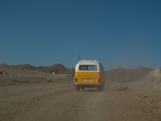 A vintage yellow and white van travels down a dusty, rugged road in a barren, mountainous landscape under a clear blue sky. Dust trails behind the vehicle, accentuating the rustic and adventurous setting.