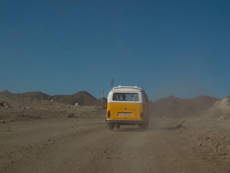 A vintage yellow and white van travels down a dusty, rugged road in a barren, mountainous landscape under a clear blue sky. Dust trails behind the vehicle, accentuating the rustic and adventurous setting.
