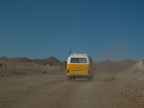 A vintage yellow and white van travels down a dusty, rugged road in a barren, mountainous landscape under a clear blue sky. Dust trails behind the vehicle, accentuating the rustic and adventurous setting.