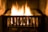A metal fireplace grate holds a burning fire with bright, yellow-orange flames casting a warm glow. The stone hearth surrounding the fire adds to the cozy ambiance.