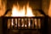 A metal fireplace grate holds a burning fire with bright, yellow-orange flames casting a warm glow. The stone hearth surrounding the fire adds to the cozy ambiance.