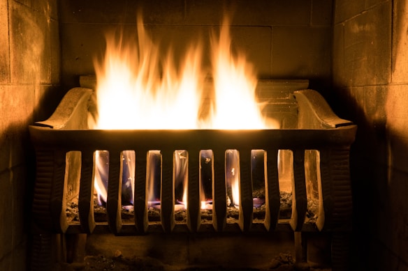 A metal fireplace grate holds a burning fire with bright, yellow-orange flames casting a warm glow. The stone hearth surrounding the fire adds to the cozy ambiance.
