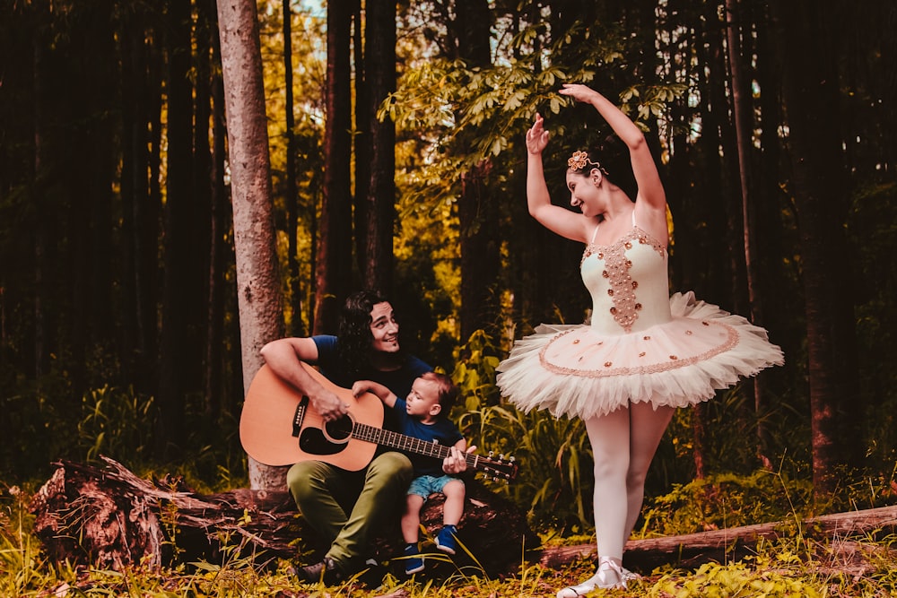 man sitting beside toddler boy while playing guitar and looking at the  woman in ballerina dress in the woods photo – Free Ballerina Image on  Unsplash