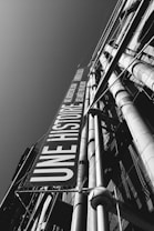 A black and white photograph captures the side of a modern building with large pipes and a prominent vertical banner. The banner reads 'UNE HISTOIRE' and stretches from the top to the bottom of the frame. The composition accentuates the industrial aesthetic with strong lines and contrasts.