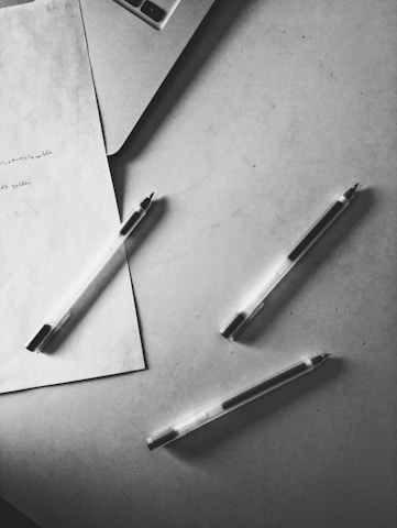 On a flat surface, three ballpoint pens are scattered around a sheet of paper with handwritten notes. A corner of a laptop is visible near the top left.
