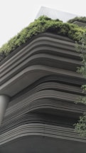 A modern building showcases layered architecture with horizontal lines creating a tiered effect. The upper levels are adorned with lush greenery, giving a natural contrast to the otherwise industrial look. The design integrates the green elements seamlessly with the structure, emphasizing eco-friendliness.