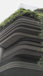 A modern building showcases layered architecture with horizontal lines creating a tiered effect. The upper levels are adorned with lush greenery, giving a natural contrast to the otherwise industrial look. The design integrates the green elements seamlessly with the structure, emphasizing eco-friendliness.