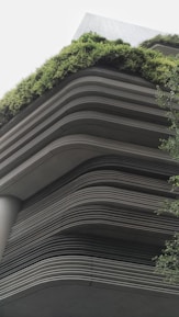 A modern building showcases layered architecture with horizontal lines creating a tiered effect. The upper levels are adorned with lush greenery, giving a natural contrast to the otherwise industrial look. The design integrates the green elements seamlessly with the structure, emphasizing eco-friendliness.