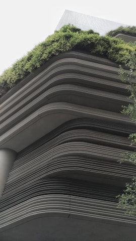 A modern building showcases layered architecture with horizontal lines creating a tiered effect. The upper levels are adorned with lush greenery, giving a natural contrast to the otherwise industrial look. The design integrates the green elements seamlessly with the structure, emphasizing eco-friendliness.