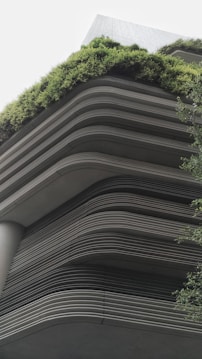 A modern building showcases layered architecture with horizontal lines creating a tiered effect. The upper levels are adorned with lush greenery, giving a natural contrast to the otherwise industrial look. The design integrates the green elements seamlessly with the structure, emphasizing eco-friendliness.