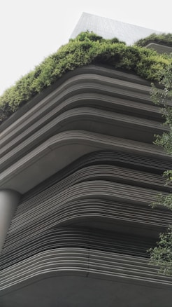 A modern building showcases layered architecture with horizontal lines creating a tiered effect. The upper levels are adorned with lush greenery, giving a natural contrast to the otherwise industrial look. The design integrates the green elements seamlessly with the structure, emphasizing eco-friendliness.