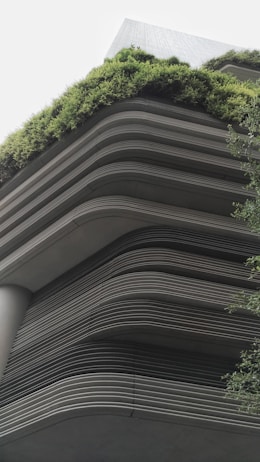 A modern building showcases layered architecture with horizontal lines creating a tiered effect. The upper levels are adorned with lush greenery, giving a natural contrast to the otherwise industrial look. The design integrates the green elements seamlessly with the structure, emphasizing eco-friendliness.