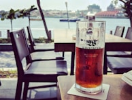A half-full glass of amber beer sits on a coaster on an outdoor table, with a scenic view of a river and buildings in the background. The scene includes empty chairs around, suggesting a relaxed, open-air dining area.