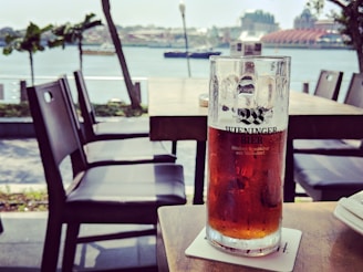 A half-full glass of amber beer sits on a coaster on an outdoor table, with a scenic view of a river and buildings in the background. The scene includes empty chairs around, suggesting a relaxed, open-air dining area.