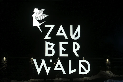 A fairy silhouette with wings and an extended arm is positioned above the stylized letters that spell out 'ZAUBERWALD'. The text and image are bright and white against a dark background. Small lights at the bottom illuminate the scene.