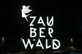 A fairy silhouette with wings and an extended arm is positioned above the stylized letters that spell out 'ZAUBERWALD'. The text and image are bright and white against a dark background. Small lights at the bottom illuminate the scene.