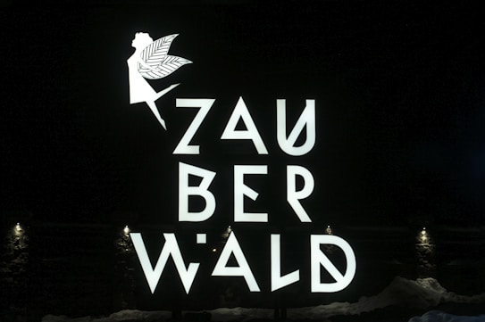 A fairy silhouette with wings and an extended arm is positioned above the stylized letters that spell out 'ZAUBERWALD'. The text and image are bright and white against a dark background. Small lights at the bottom illuminate the scene.