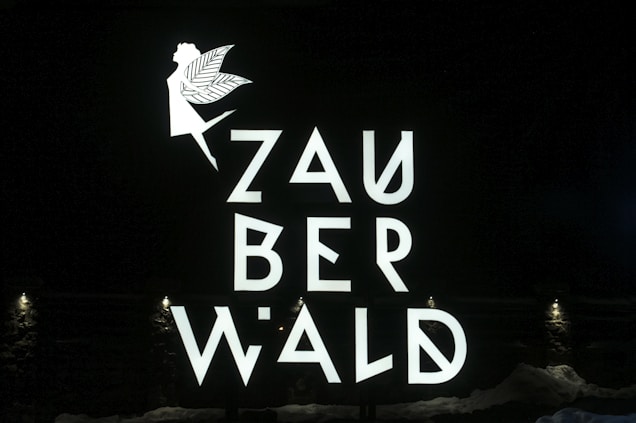 A fairy silhouette with wings and an extended arm is positioned above the stylized letters that spell out 'ZAUBERWALD'. The text and image are bright and white against a dark background. Small lights at the bottom illuminate the scene.