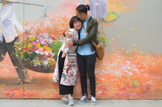 A young couple is embracing affectionately in front of a colorful mural depicting a vendor with a basket of vibrant flowers. The mural has a mix of autumn colors with falling leaves, enhancing the romantic atmosphere. The couple seems to be enjoying their moment, smiling and displaying a relaxed, joyful disposition.