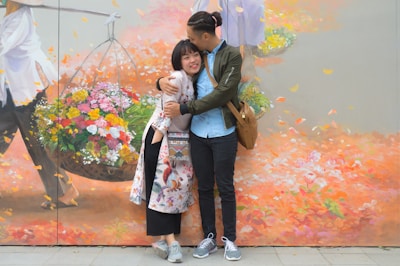 A young couple is embracing affectionately in front of a colorful mural depicting a vendor with a basket of vibrant flowers. The mural has a mix of autumn colors with falling leaves, enhancing the romantic atmosphere. The couple seems to be enjoying their moment, smiling and displaying a relaxed, joyful disposition.