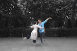 A couple is dancing playfully on a paved surface in an outdoor setting surrounded by dense foliage. The woman is wearing a long, white floral dress and has one leg lifted in the air, while the man wears a blue shirt with a camera hanging around his neck and is holding her, extending his other arm outwards.