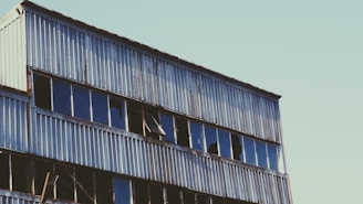 A dilapidated industrial building with rusted metal siding and broken windows. The structure appears neglected and abandoned, with glass panes shattered and missing.