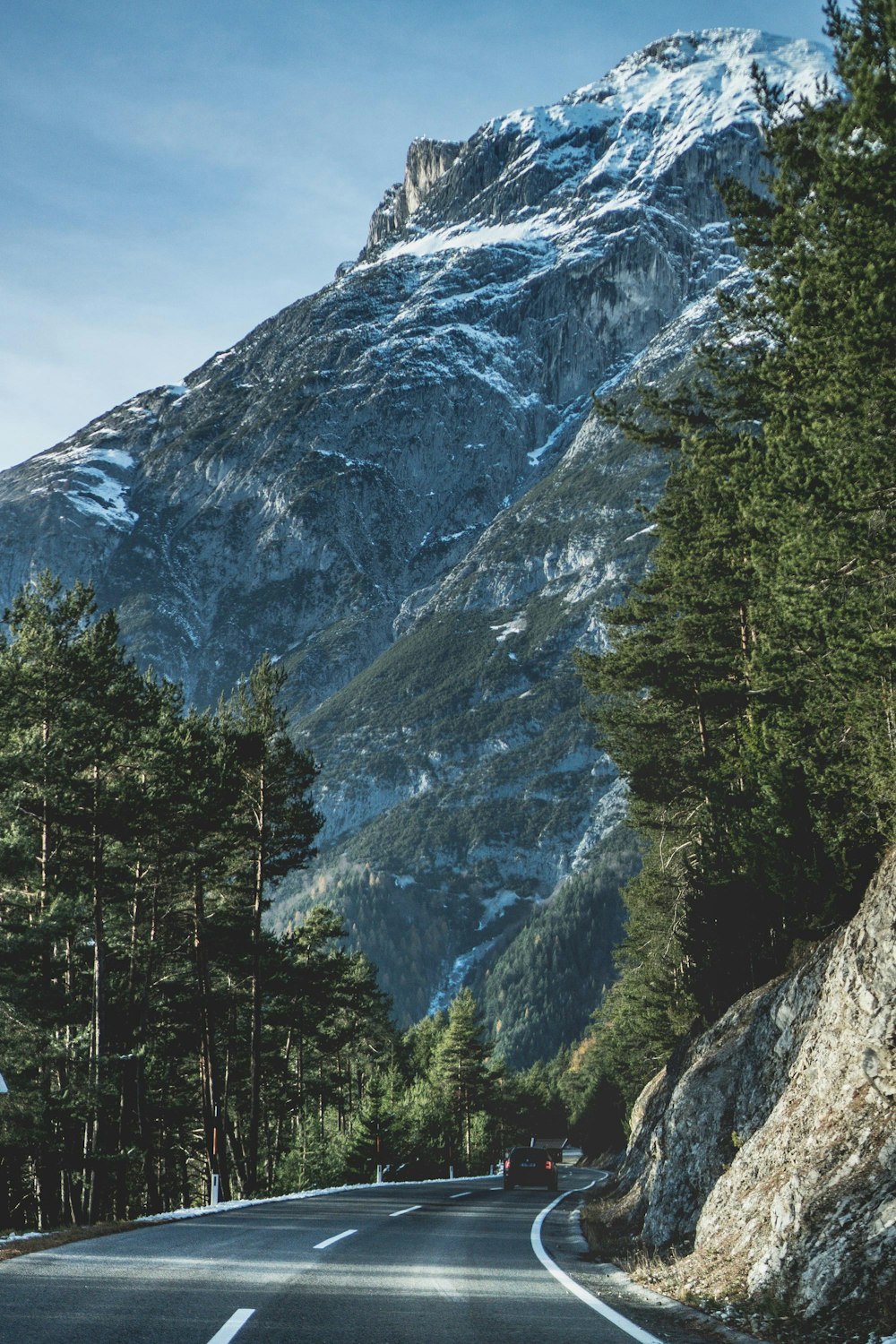 Photographie de route en béton entre les arbres et la falaise avec fond de montagne