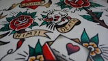 A vibrant image featuring classic tattoo art with elements such as a rose, skull, heart, and banners with text like 'AMORE', 'TIL DEATH', and 'HIM?!'. The design includes bold outlines and colors, resembling traditional tattoo flash sheets.