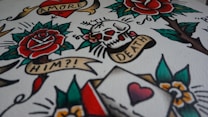 A vibrant image featuring classic tattoo art with elements such as a rose, skull, heart, and banners with text like 'AMORE', 'TIL DEATH', and 'HIM?!'. The design includes bold outlines and colors, resembling traditional tattoo flash sheets.
