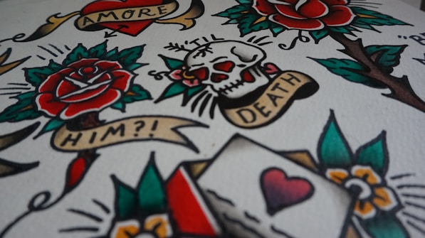 A vibrant image featuring classic tattoo art with elements such as a rose, skull, heart, and banners with text like 'AMORE', 'TIL DEATH', and 'HIM?!'. The design includes bold outlines and colors, resembling traditional tattoo flash sheets.