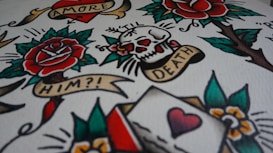 A vibrant image featuring classic tattoo art with elements such as a rose, skull, heart, and banners with text like 'AMORE', 'TIL DEATH', and 'HIM?!'. The design includes bold outlines and colors, resembling traditional tattoo flash sheets.