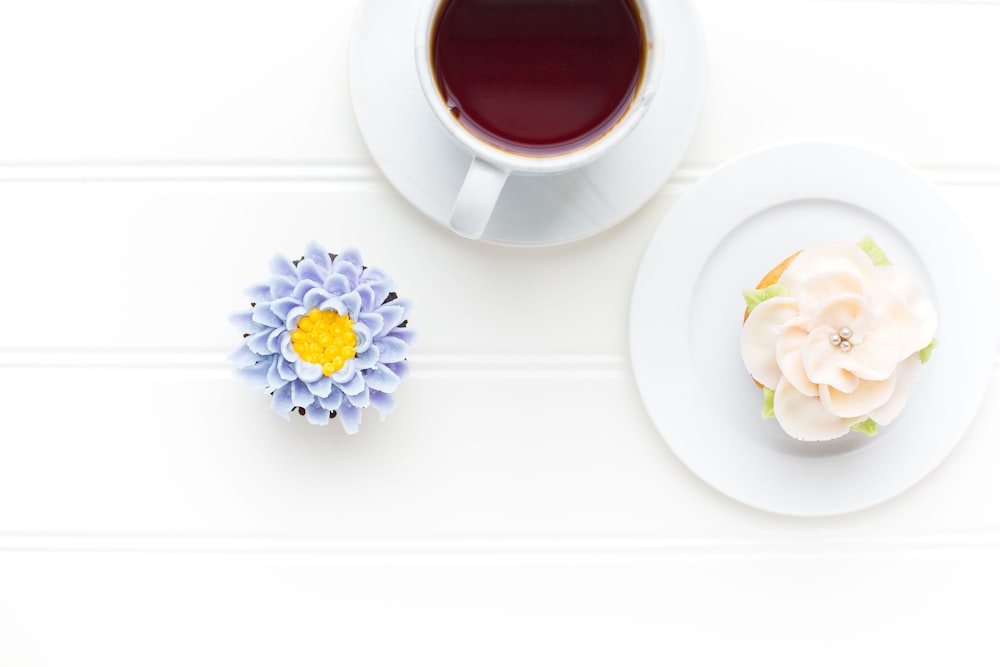 Fotografía plana de taza de café blanco en plato de platillo blanco y flores púrpuras