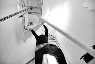 A person is installing or adjusting a folding attic ladder. The scene is captured from a low angle, emphasizing the structure of the ladder and the ceiling. The workspace is well-lit with light reflecting off the walls and ceiling, creating a high-contrast environment.