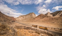A railway winds through a rugged, mountainous desert landscape under a bright blue sky with patches of white clouds. The terrain is dry with scattered shrubs and grass, surrounded by steep rock formations.