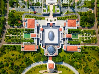 An aerial view of a large, symmetrical building complex surrounded by lush greenery. The structure features red-tiled roofs and geometric patterns with two prominent dome-shaped glass ceilings. The grounds include neatly arranged gardens, pathways, and parking areas.