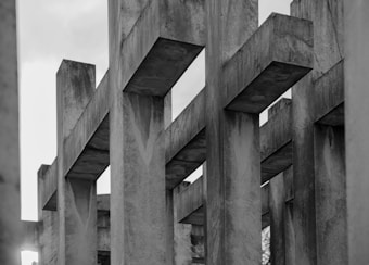 A series of large, rectangular concrete beams arranged in a geometric pattern, creating a sense of depth and perspective. The structure is composed of repetitive vertical and horizontal lines that converge in the center. The surface of the concrete appears weathered, with visible textures and stains.