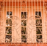An intricately designed wrought iron grille covers a window, featuring swirling and circular patterns. The grille is painted in a warm, pastel shade, complementing the surrounding architecture. Behind the grill, a framed window with clear glass reflects some greenery.