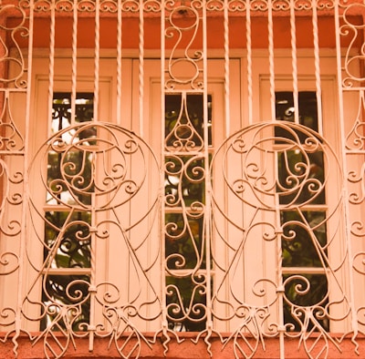 An intricately designed wrought iron grille covers a window, featuring swirling and circular patterns. The grille is painted in a warm, pastel shade, complementing the surrounding architecture. Behind the grill, a framed window with clear glass reflects some greenery.