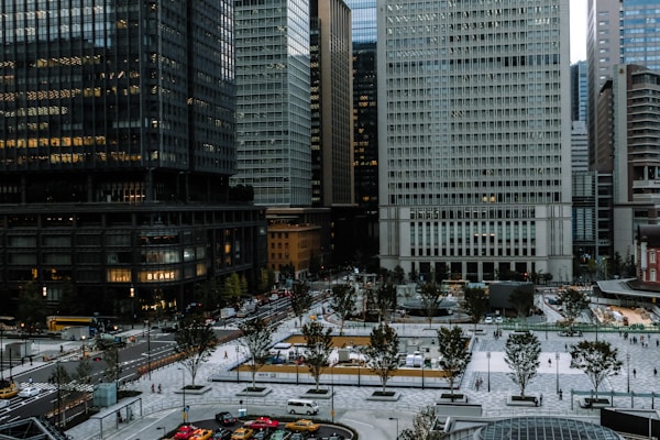 A bustling urban setting featuring high-rise buildings with reflective glass surfaces. The foreground includes a spacious paved plaza with scattered trees and people walking. Traffic is visible on the roads, with several cars and buses navigating the area. The architecture is modern, and the environment appears densely populated.