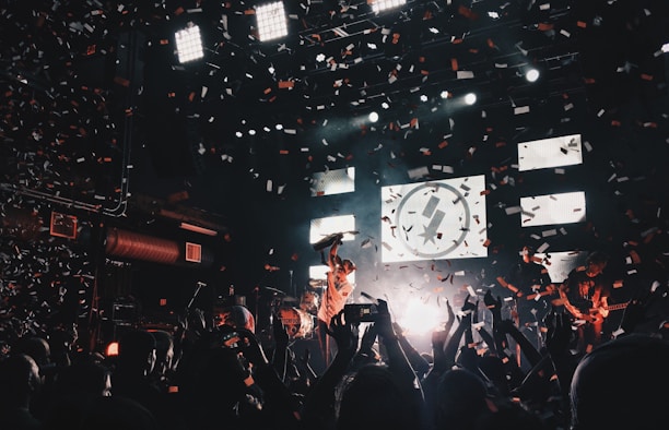 A lively concert scene with a band performing on stage, surrounded by an enthusiastic crowd. Confetti fills the air as bright stage lights illuminate the musicians and the audience. The atmosphere is vibrant, with people capturing the moment on their phones.