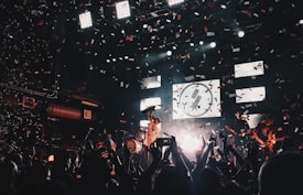 A lively concert scene with a band performing on stage, surrounded by an enthusiastic crowd. Confetti fills the air as bright stage lights illuminate the musicians and the audience. The atmosphere is vibrant, with people capturing the moment on their phones.