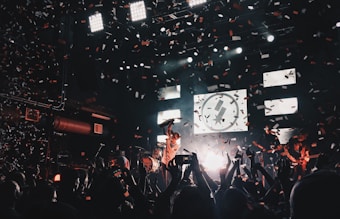 A lively concert scene with a band performing on stage, surrounded by an enthusiastic crowd. Confetti fills the air as bright stage lights illuminate the musicians and the audience. The atmosphere is vibrant, with people capturing the moment on their phones.