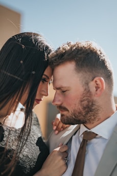 A close-up of two people, a woman and a man, standing intimately close with their foreheads touching. The woman has long dark hair and is wearing a black outfit, while the man has short hair and a beard, wearing a light-colored suit and a brown tie. The background is softly blurred, suggesting an outdoor setting with a clear sky.