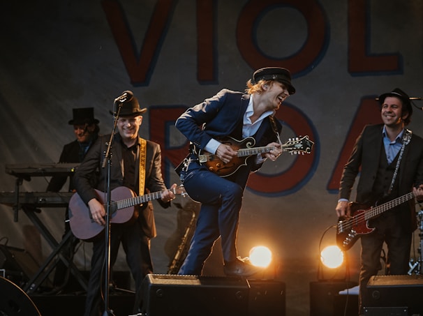 A band performing on stage with four musicians. The central figure, wearing a hat, energetically plays a mandolin. The others play various instruments including a guitar and a keyboard. Stage lighting creates a lively atmosphere as they smile and engage with the performance.