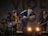 A band performing on stage with four musicians. The central figure, wearing a hat, energetically plays a mandolin. The others play various instruments including a guitar and a keyboard. Stage lighting creates a lively atmosphere as they smile and engage with the performance.