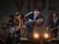 A band performing on stage with four musicians. The central figure, wearing a hat, energetically plays a mandolin. The others play various instruments including a guitar and a keyboard. Stage lighting creates a lively atmosphere as they smile and engage with the performance.