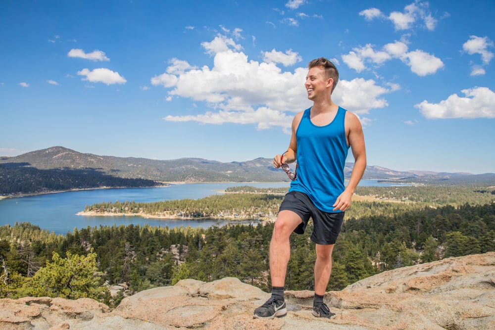 Mann mit blauem Tanktop und schwarzen Shorts steht auf Felsklippe