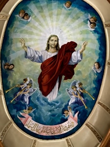 A religious painting featuring a central figure with raised arms, surrounded by a radiant glow. The figure is clothed in a red and white robe, with angels and cherubs positioned in the corners and along the edges. Trumpet-holding angels are prominently featured, emerging from a backdrop of soft, cloud-like formations. An inscription on a ribbon floats at the bottom, written in an ornate script.