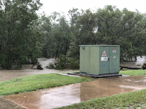 A green electrical box with warning signs sits on a small concrete platform next to a lush patch of grass. Behind it, a swollen river flows amidst numerous green trees, suggesting a recent heavy rain or flooding. The sky appears overcast, and the ground is wet, reflecting the recent or ongoing rainfall.