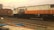 A freight train with a locomotive painted in white, orange, and blue, pulling a cargo container. The train is on a set of railway tracks with electric lines overhead and some industrial elements in the background. A few cables and parts of a motorcycle are visible in the foreground.